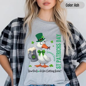 A light grey t-shirt featuring a cartoon goose dressed for St. Patricks Day with a green bow tie, leprechaun hat, and shamrocks. The goose, named Guinness Goose, holds a frothy beer mug labeled GG with a playful, mischievous expression. Surrounding elements include shamrocks. The words ST PATRICKS DAY are written vertically on the right, with the words How the Heck Am I Getting Home? are written horizontally below.