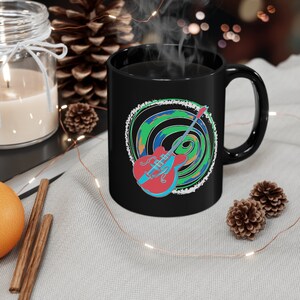May include: Black coffee mug with a colorful guitar graphic and a swirling green, blue, and white design. The guitar is red, blue, and black.