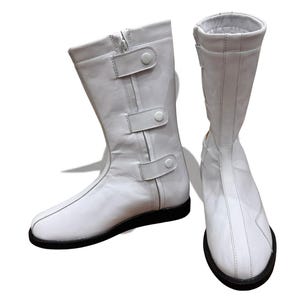 Puede incluir: Un par de botas de cuero blanco con suelas negras y hebillas. Las botas tienen una cremallera en el interior de cada bota.