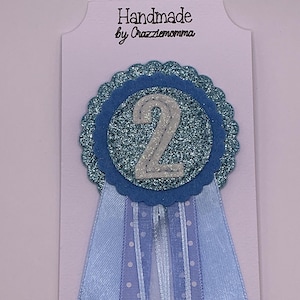May include: A handmade blue rosette with a glittery center and the number 2 in white. The rosette is attached to a light blue ribbon with polka dots and the text "Handmade by Crazzlemomma".