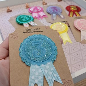 Puede incluir: Insignia artesanal azul brillante con el número tres, borde festoneado y cinta de lunares. Fijada a una tarjeta marrón con el texto "Handmade by Crazzlemomma". Otras insignias coloridas son visibles en el fondo.