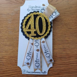 May include: A black and gold glitter 40th birthday badge with three white ribbons that say "Happy 40th" with a gold ribbon and a gold bow.