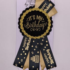 May include: A black and gold birthday rosette with the text "IT'S MY Birthday" in gold lettering. The rosette has gold glitter trim and ribbons that say "Happy Birthday". A cake graphic is also visible.