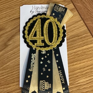 May include: A handmade 40th birthday badge with a black and gold color scheme. The badge features a black scalloped circle with a gold glitter "40" in the center. Black ribbons with gold text and star accents hang below, with the words "Happy Birthday" and "Happy".