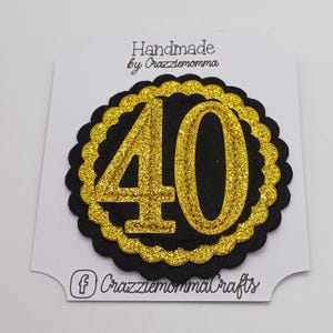 May include: A black and gold glittery "40" birthday decoration. The number "40" is in gold glitter, set against a black scalloped circle, with a gold glitter border. The item is on a white card with the text "Handmade by Crazziemomma" and a social media icon.