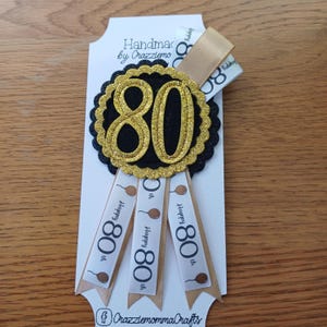 May include: An 80th birthday badge featuring a black circle with a gold glitter number 80 and a scalloped edge. The badge is attached to three white ribbons with gold trim and the text "Happy 80th" printed on each ribbon.