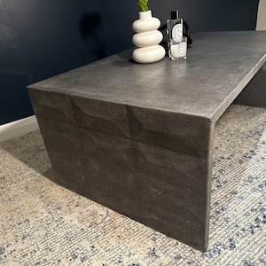May include: A dark grey concrete coffee table with a unique geometric design on the front. The table has a rectangular top and a wide, open base.