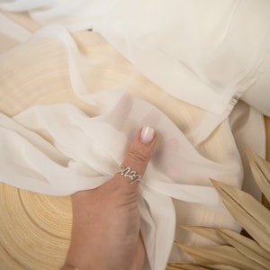 May include: White sheer fabric rolled up on a wooden surface. The fabric is very thin and delicate, and it has a slight sheen. The fabric is being held by a hand wearing a ring.
