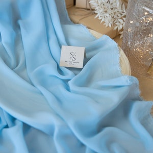 May include: A light blue fabric is draped, with soft folds and a frayed edge. A small card with the text "SECRET SPARK fabrics" is visible. The background includes wrapped packages and a decorative vase.