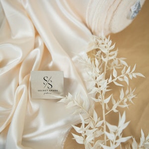 May include: A close-up of a cream-coloured fabric roll with a white label. The fabric is folded and rolled up. The label has the text "SECRET SPARK fabrics" on it. There is a small square card with the text "SECRET SPARK fabrics" and a logo of two overlapping "S" letters. The card is on top of a cream-coloured satin fabric. A white dried flower branch is on the right side of the image.