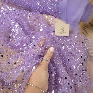 May include: Purple sequin fabric with a sheer mesh backing. The fabric is covered in small, round sequins that are scattered randomly across the surface. The fabric is soft and delicate, and it would be perfect for a variety of projects, such as dresses, skirts, and tops.