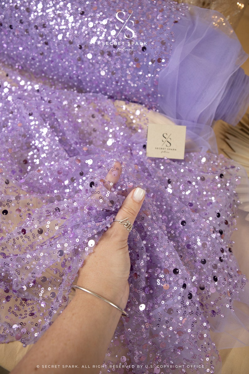 May include: Purple sequin fabric with a sheer mesh backing. The fabric is covered in small, round sequins that are scattered randomly across the surface. The fabric is soft and delicate, and it would be perfect for a variety of projects, such as dresses, skirts, and tops.