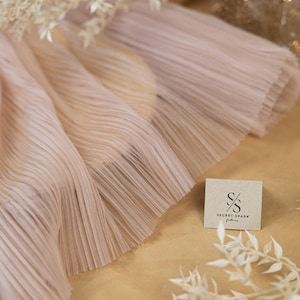 May include: Close-up of a pale pink pleated fabric with a delicate, ruffled edge. The fabric is soft and flowing, with a subtle sheen. The fabric is displayed on a brown surface with a small, white tag that reads "SECRET SPARK fabrics".