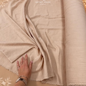 May include: A roll of light beige fabric with a soft, crinkled texture. The fabric is partially unrolled, revealing a raw, frayed edge. The logo for Secret Spark Fabrics is visible at the top, and text at the bottom reads Secret Spark.