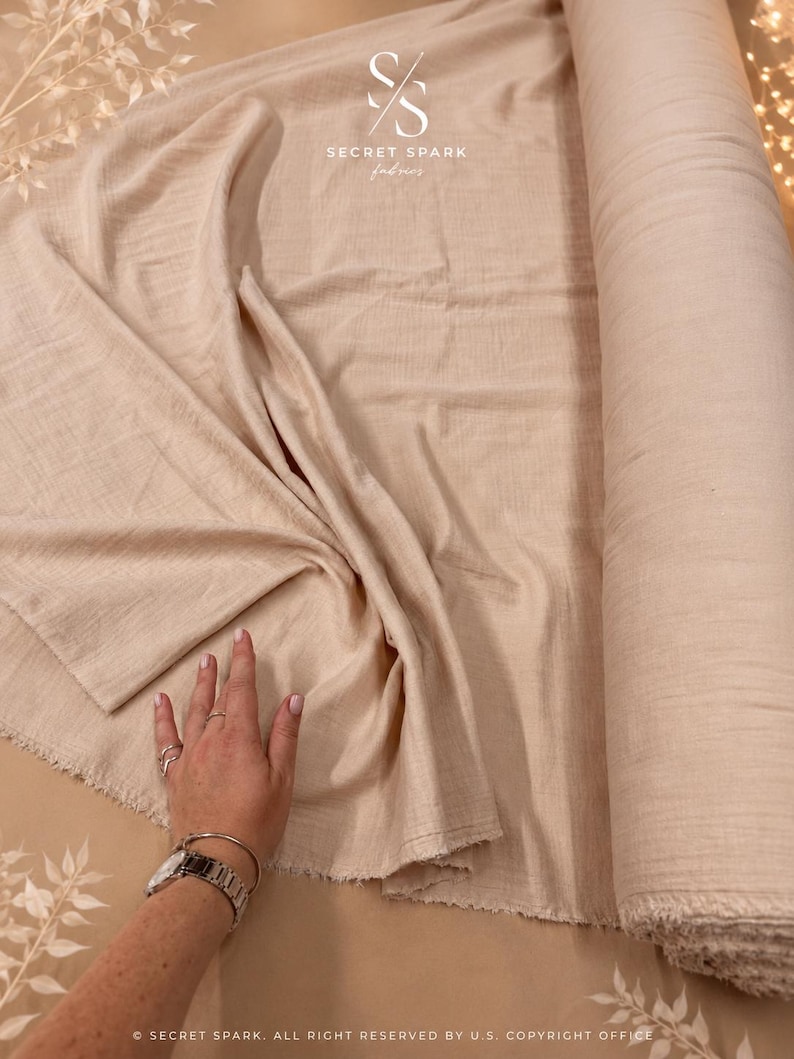 May include: A roll of light beige fabric with a soft, crinkled texture. The fabric is partially unrolled, revealing a raw, frayed edge. The logo for Secret Spark Fabrics is visible at the top, and text at the bottom reads Secret Spark.