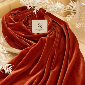 Puede incluir: Un primer plano de una tela de terciopelo de color óxido drapeada sobre una superficie de madera. La tela es lisa y tiene un brillo sutil. La tela está etiquetada con el texto "SECRET SPARK" en letras negras.