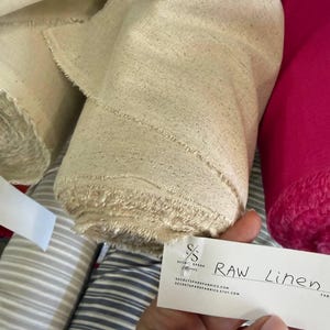 May include: A roll of natural beige raw linen fabric with a visible woven texture and frayed edges. A white rectangular tag is attached, featuring the text "SS SECRET SPARK FABRICS" and "RAW Linen" along with the website address and Etsy shop URL.