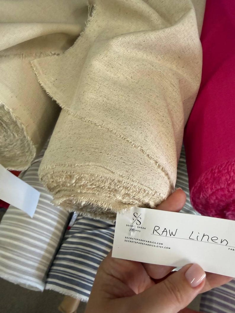 May include: A roll of natural beige raw linen fabric with a visible woven texture and frayed edges. A white rectangular tag is attached, featuring the text "SS SECRET SPARK FABRICS" and "RAW Linen" along with the website address and Etsy shop URL.