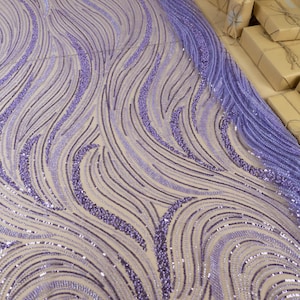 May include: Close-up of a fabric with a wave-like pattern in shades of lavender and purple. The fabric is embellished with sequins and beads, creating a shimmering effect. The text "SECRET SPARK fabrics" is visible.