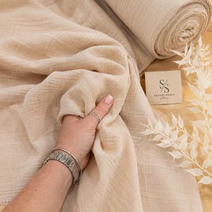 May include: A roll of beige muslin fabric with a crinkled texture, featuring a square tag with the text Secret Spark and a logo consisting of the letters S and S. The fabric has a raw, frayed edge and a soft, lightweight appearance.