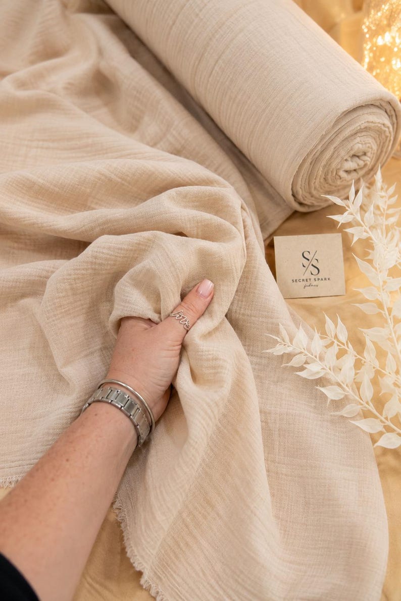 May include: A roll of beige muslin fabric with a crinkled texture, featuring a square tag with the text Secret Spark and a logo consisting of the letters S and S. The fabric has a raw, frayed edge and a soft, lightweight appearance.