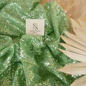 May include: Green sequin fabric with a light green mesh backing. The fabric is draped over a wooden surface and a clear glass vase. The fabric has a small card on top with the text "SECRET SPARK fabrics" and a logo of two overlapping "S" letters.