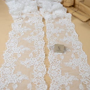 Puede incluir: Dos tiras de tela de encaje floral blanco se muestran sobre una superficie beige. El encaje presenta intrincados patrones florales y un borde festoneado. La marca "SECRET SPARK fabrics" es visible.