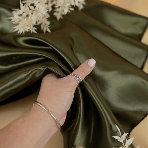 May include: A close-up of a hand holding a green satin fabric. The fabric is smooth and silky. The hand is wearing a silver ring with the word "love" on it. The fabric is draped over a table with a white flower arrangement in the background.