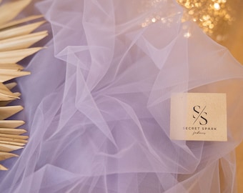 May include: A close-up of a light purple tulle fabric draped over a wooden circle. The fabric is very sheer and delicate. The fabric is labeled with the text "SECRET SPARK fabrics".