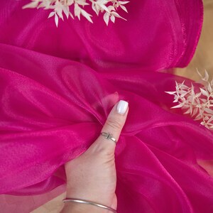 May include: A bright pink sheer fabric with a slight shimmer. The fabric is draped over a hand and is being held in place by a finger.