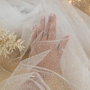 May include: A sheer, white fabric with a delicate, dotted pattern. The fabric is draped, with a hand visible through the material. The image includes the text "SECRET SPARK fabrics".