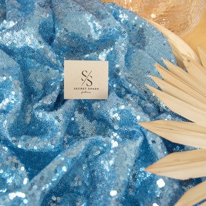 May include: A close-up of a light blue sequined fabric with a small brown card that says "SECRET SPARK fabrics" with the initials "SS" in a circle.
