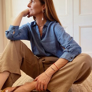 May include: A blue long-sleeved button-down shirt with a collar, paired with loose-fitting tan trousers. The outfit is accessorized with gold hoop earrings featuring dangling white pearls, layered necklaces, a thin red bracelet, and brown thong sandals.