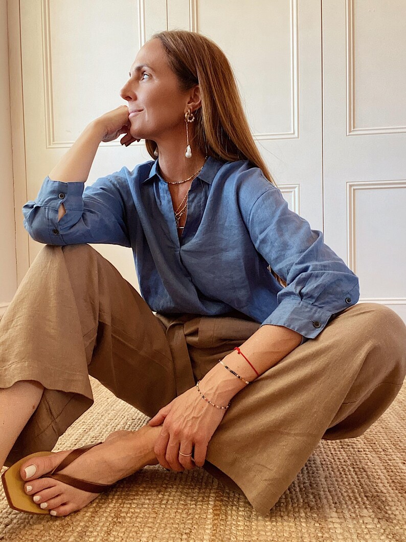 May include: A blue long-sleeved button-down shirt with a collar, paired with loose-fitting tan trousers. The outfit is accessorized with gold hoop earrings featuring dangling white pearls, layered necklaces, a thin red bracelet, and brown thong sandals.