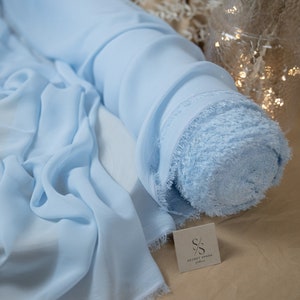 Pode incluir: Um rolo de tecido de chiffon azul claro com uma borda franjada. O tecido est&aacute; enrolado e amarrado com uma fita branca. O tecido est&aacute; sobre uma superf&iacute;cie marrom com uma etiqueta branca que diz "SECRET SPARK fabrics".