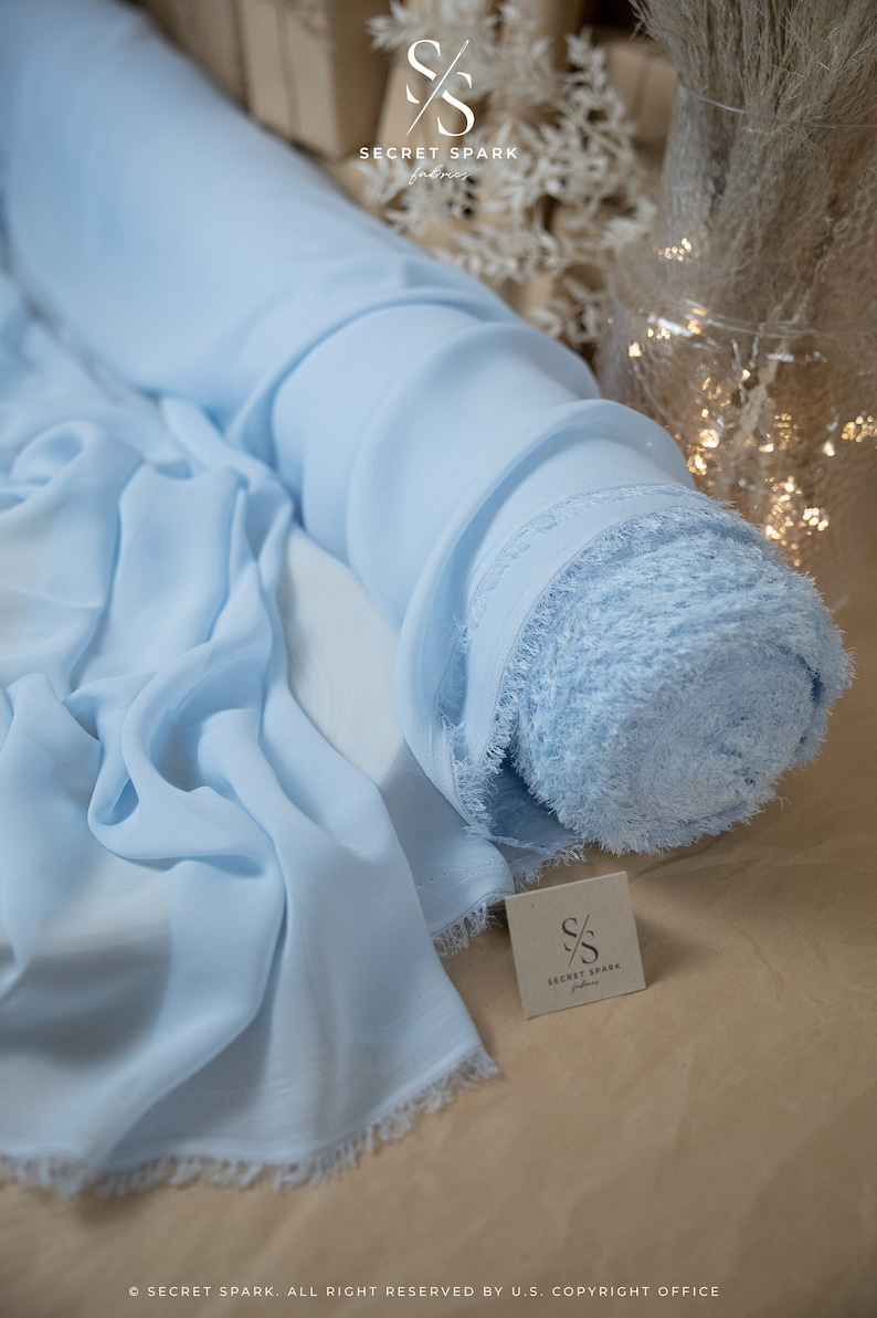 Pode incluir: Um rolo de tecido de chiffon azul claro com uma borda franjada. O tecido est&aacute; enrolado e amarrado com uma fita branca. O tecido est&aacute; sobre uma superf&iacute;cie marrom com uma etiqueta branca que diz "SECRET SPARK fabrics".