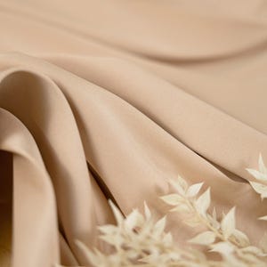 May include: Close-up of a light brown fabric with a subtle texture. The fabric is draped in soft folds, creating a luxurious and elegant look. The fabric is highlighted by a sprig of dried white flowers.