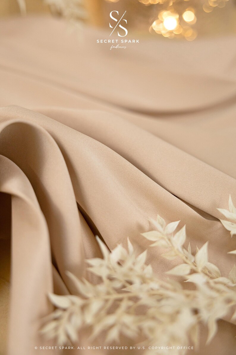 May include: Close-up of a light brown fabric with a subtle texture. The fabric is draped in soft folds, creating a luxurious and elegant look. The fabric is highlighted by a sprig of dried white flowers.