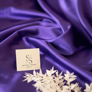 Pu&ograve; includere: Primo piano di un tessuto di raso viola con un ramoscello di fiori secchi bianchi e un piccolo biglietto con il testo "Secret Spark Fabrics" e il logo "SS".