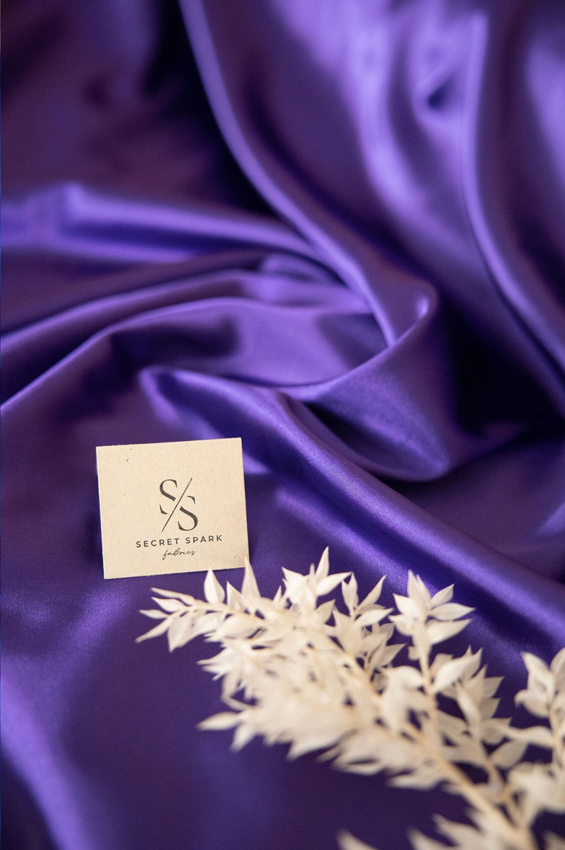 Pu&ograve; includere: Primo piano di un tessuto di raso viola con un ramoscello di fiori secchi bianchi e un piccolo biglietto con il testo "Secret Spark Fabrics" e il logo "SS".