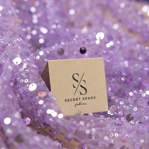 May include: A close-up of a brown card with the text "SECRET SPARK fabrics" and a stylized "SS" logo on a purple sequined fabric.
