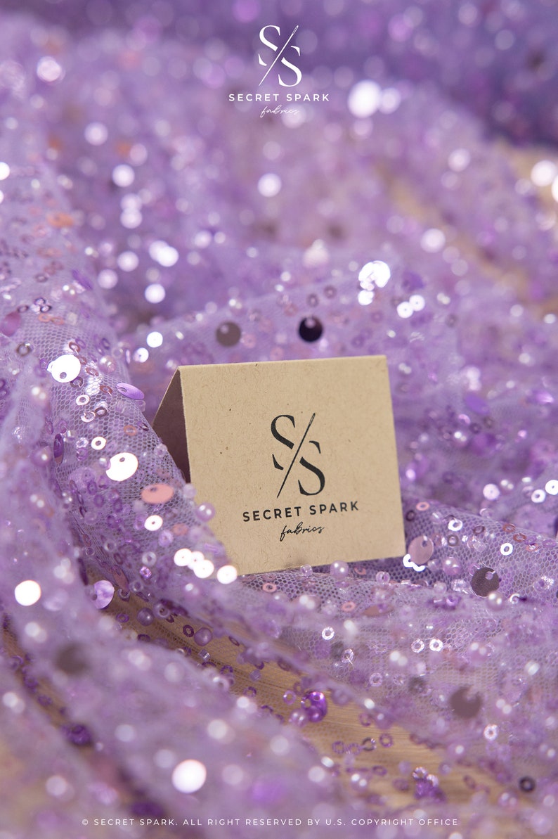 May include: A close-up of a brown card with the text "SECRET SPARK fabrics" and a stylized "SS" logo on a purple sequined fabric.
