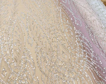 May include: Close-up of a fabric with intricate silver sequin and bead embellishments. The fabric has a beige base with a floral pattern. The text "SECRET.SPARK" is visible. Rolls of fabric in various colors are in the background.