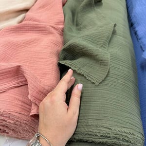 May include: Multiple rolls of soft, crinkled double gauze fabric in various solid colors including cream, dusty rose, olive green, and blue. The fabric features a distinct textured, puckered surface with raw, frayed edges along the cut ends.