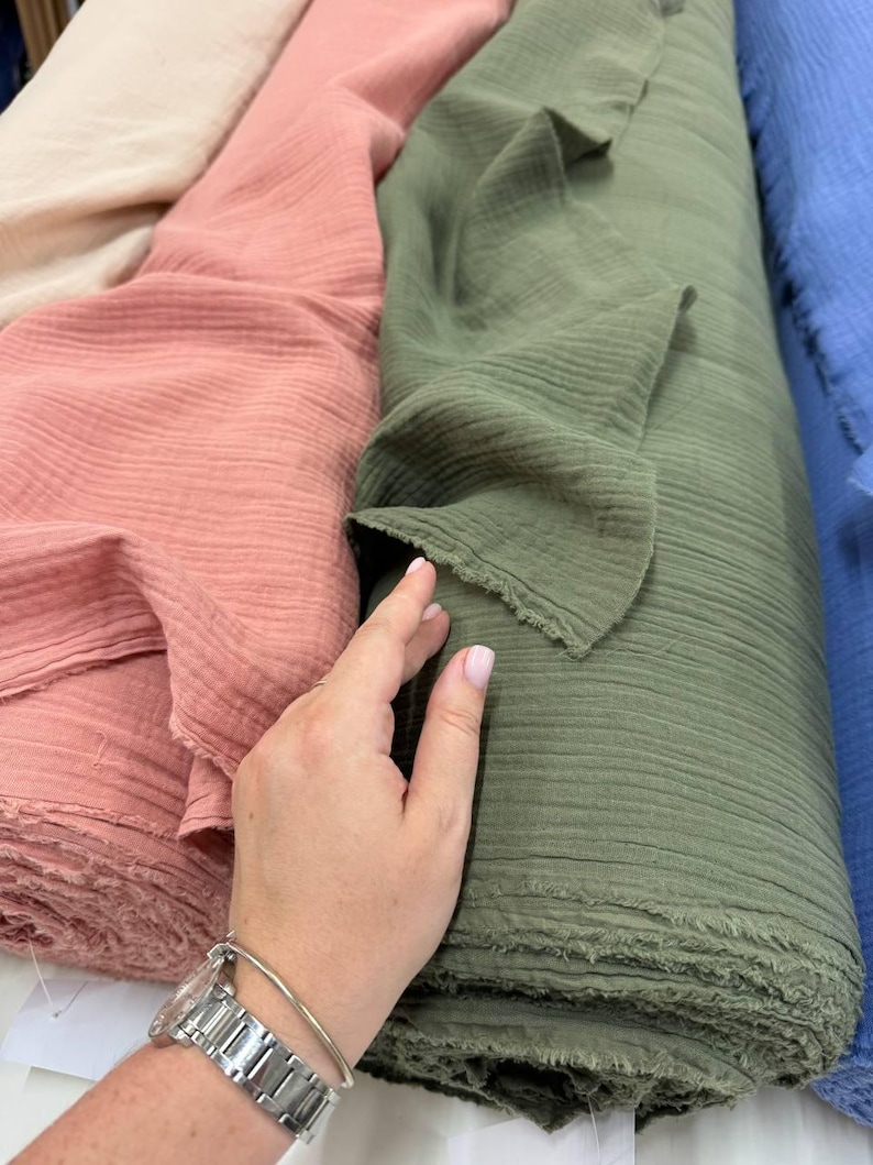 May include: Multiple rolls of soft, crinkled double gauze fabric in various solid colors including cream, dusty rose, olive green, and blue. The fabric features a distinct textured, puckered surface with raw, frayed edges along the cut ends.