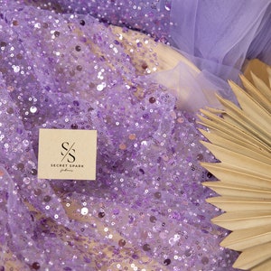 May include: Purple sequin fabric with a sheer mesh backing. The fabric is decorated with small, round sequins in a variety of sizes. The fabric is shown on a brown surface with a palm leaf fan.