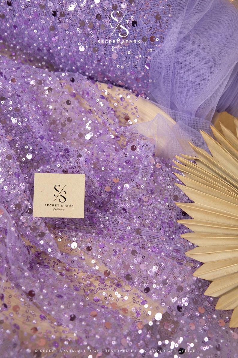 May include: Purple sequin fabric with a sheer mesh backing. The fabric is decorated with small, round sequins in a variety of sizes. The fabric is shown on a brown surface with a palm leaf fan.