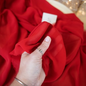 Puede incluir: Un primer plano de una tela roja transparente brillante, sostenida en una mano. La tela es suave y fluida, con una textura suave y delicada. La tela es perfecta para manualidades, costura y otros proyectos creativos.