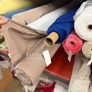 以下が含まれることがあります： 棚に保管された生地のロールのコレクション。EARTHY BROWNと印刷された目立つアースブラウンのロールが含まれています。その他、黄色、ライトベージュ、白、ロイヤルブルー、ピンク、白のストライプ柄の生地ロールが見えます。