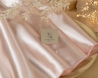 Op de afbeelding: Een licht roze satijnen stof met een subtiele glans. De stof is gevouwen en gepresenteerd op een houten bord. Een klein bruin kaartje met de tekst "SECRET SPARK fabrics" is op de stof gelegd.